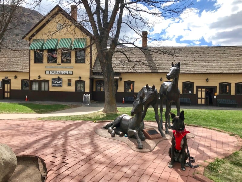 Brindle dog in a red bandana and harness sitting in front of a sculpture of three horses on Main Avenue in Durango, CO