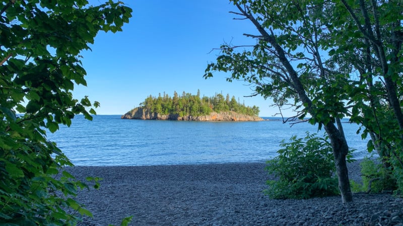 View of a wooded island on Lake Superior from a beach made of large, smooth pebbles.