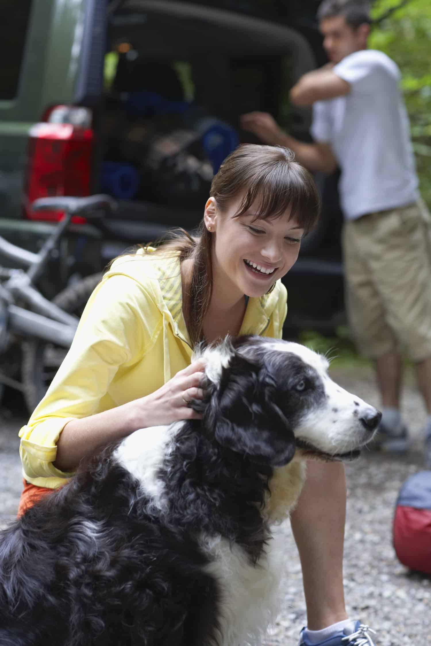 Woman with dog outdoors, man loading car in background