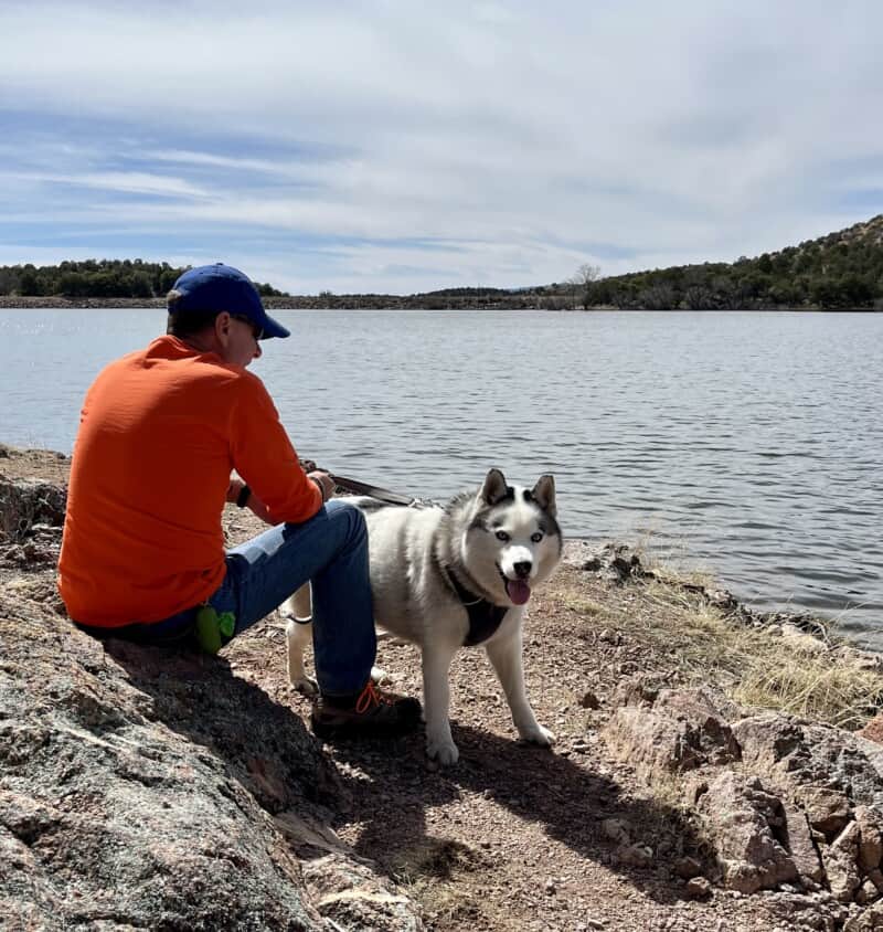 Man in an orange shirt and blue hat with a white and grey dog with arthritis on the shore of a lake
