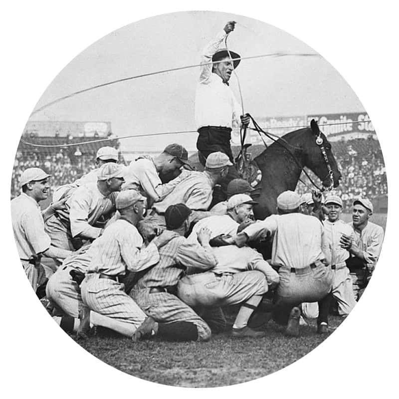 Black and white photo of Will Rogers horseback swinging a lasso around a baseball team