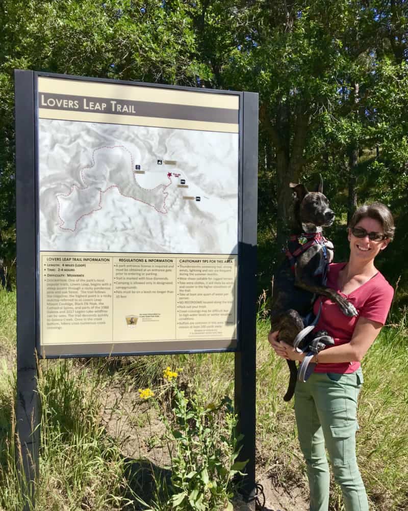 Woman holding a brindle dog beside the trailhead sign for Lover's Leap Trail in pet friendly Custer State Park, South Dakota