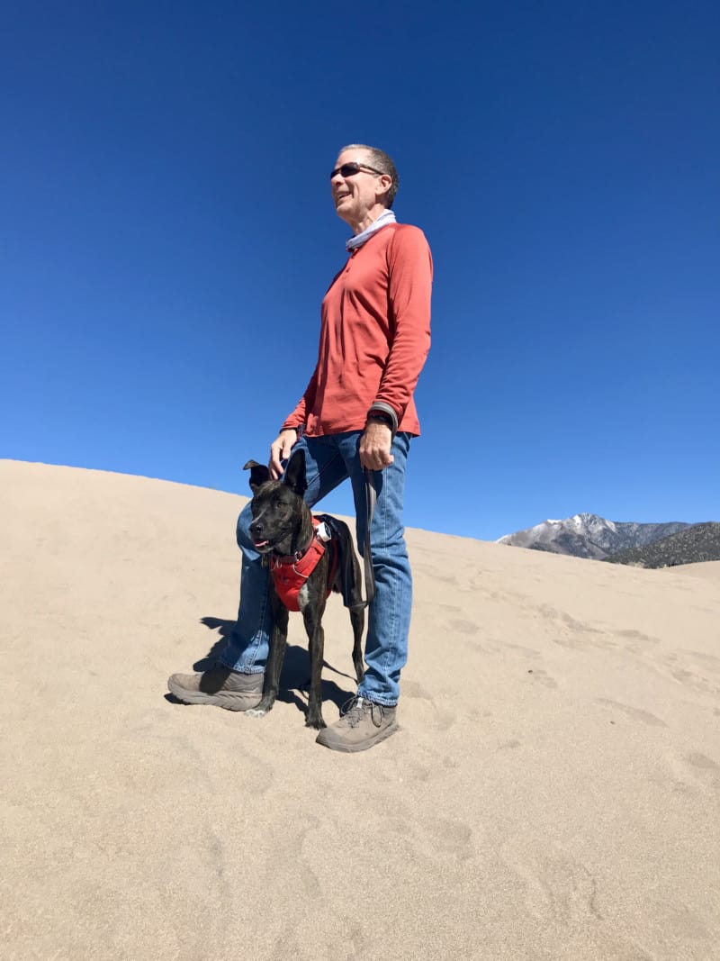 رجل يقف على الكثبان الرملية مع كلب بين ساقيه في حديقة Great Sand Dunes الوطنية