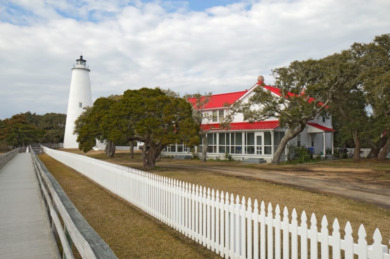 White picket fence and walkway lead past the red-roofed keepers quarters towards the tower of the Ocracoke Island lighthouse on the dog friendly Outer Banks of North Carolina