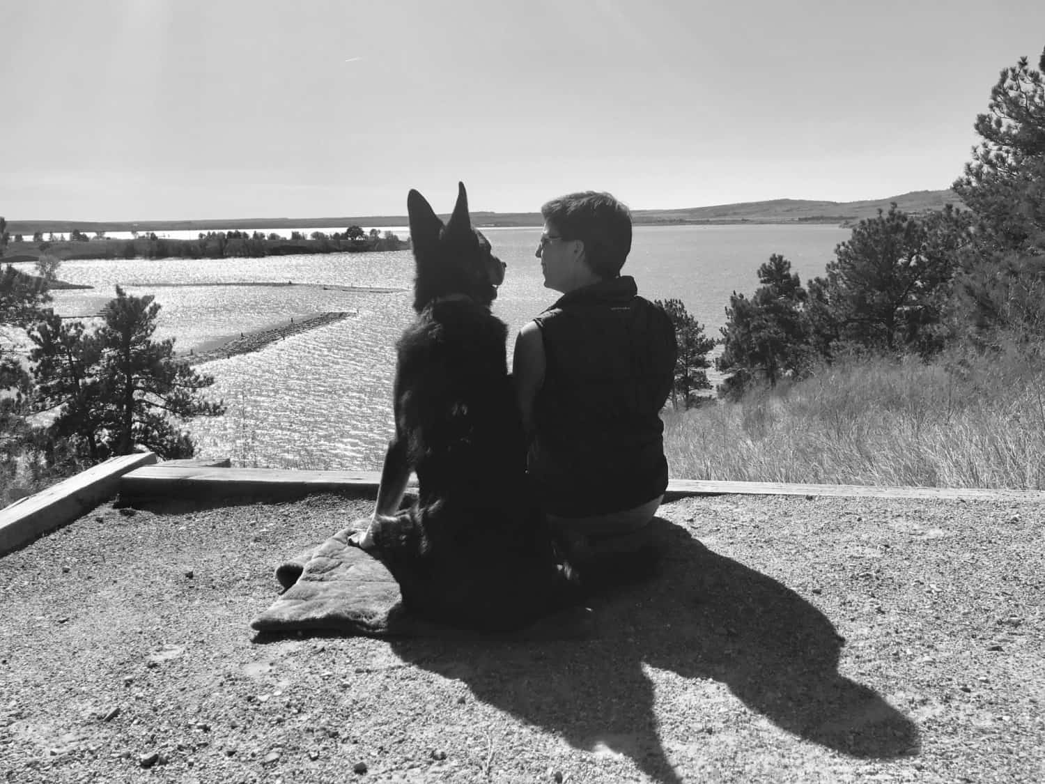 Black and white photo of a German Shepherd Dog and a woman smiling at each other overlooking a lake