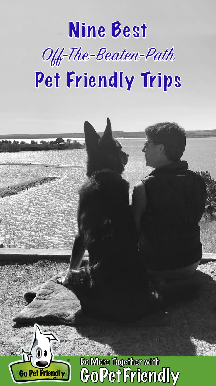 Black and white photo of a woman and a German Shepherd Dog sitting together on a bluff overlooking a lake smiling at each other