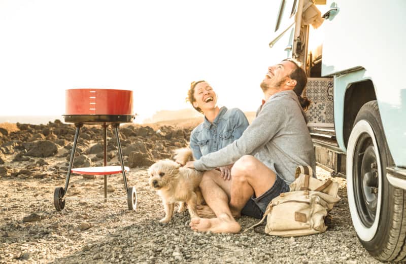 Hipster couple with cute dog traveling together on oldtimer mini van transport - Travel lifetstyle concept with indie people on minivan adventure trip having fun in barbecue moment - Warm retro filter