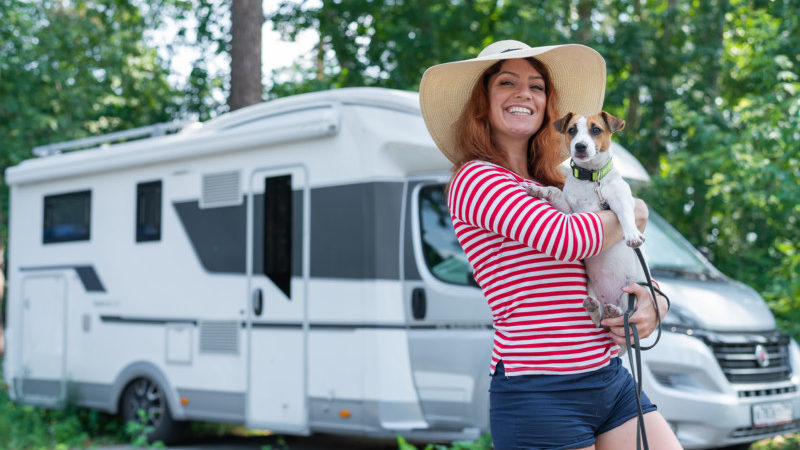 Caucasian red-haired woman in a straw hat and striped sweater walks with a small dog in front of a mobile home. Travel in a camper van with your pet.