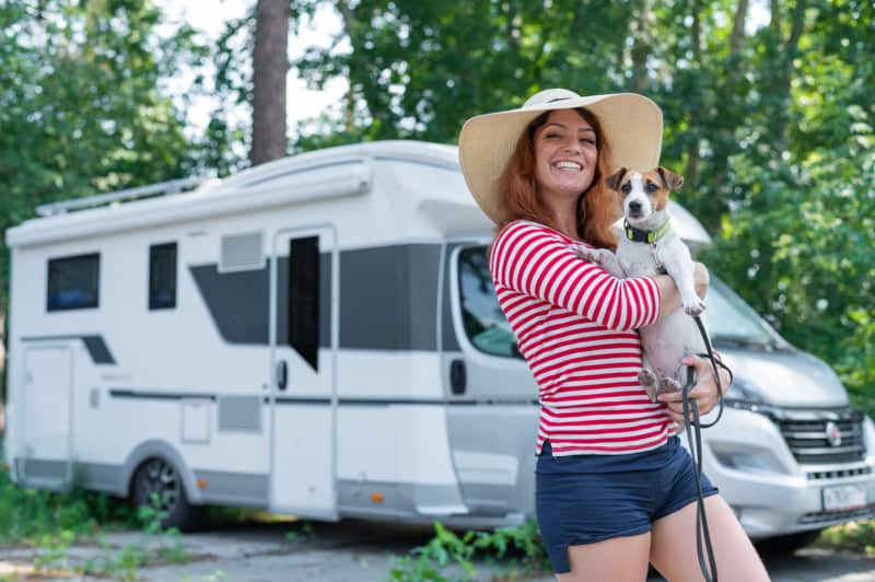 A red-haired Caucasian woman in a straw hat and striped sweater walks in front of a mobile home with a small dog. Travel in a campervan with your pet.