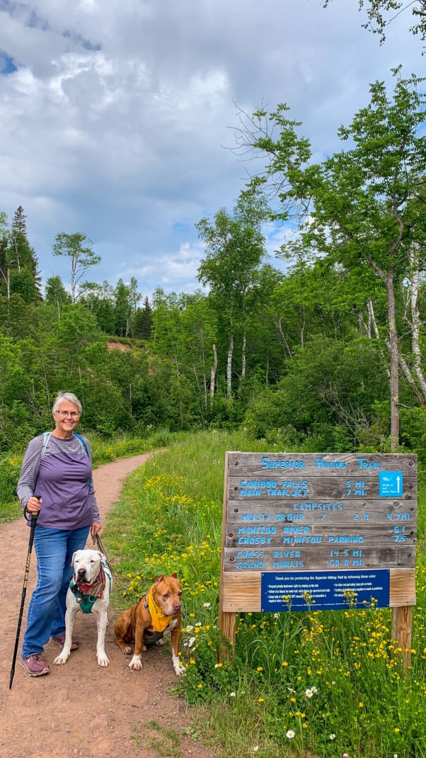 A woman, a white dog, and a brown dog standing at the start of a hiking trail next to the trail sign at Caribou Falls State Wayside on the North Shore in Minnesota.