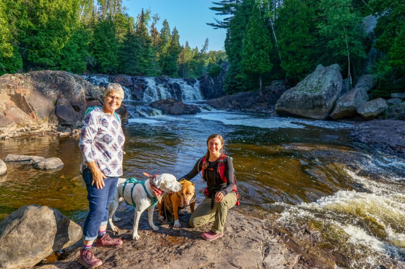 Two women and two dogs in front of a waterfall with trees in the background at Tettegouche State Park in Minnesota