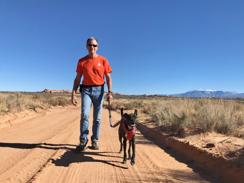 Man in orange t-shirt walking a smiling dog on Willow Flats Road in Arches National Park - Moab, UT