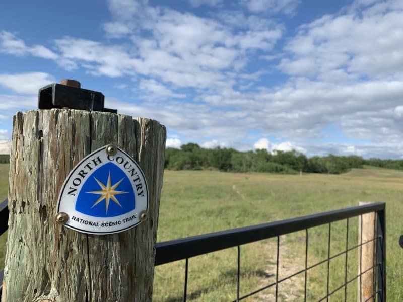 A North Country National Scenic Trail sign at a gate leading to a hiking trail in a grassy field, one of the best dog friendly spots on North Dakota