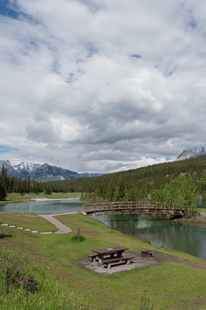 View of pet friendly picnic area in Banff. Cascade Ponds. Picnic tables, mountains and a wooden bridge over water are seen. 
