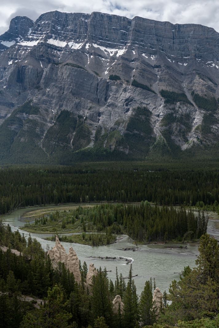 Landscape image of Banff. Hoodoos, forest, glacial river and enormous mountain are seen.