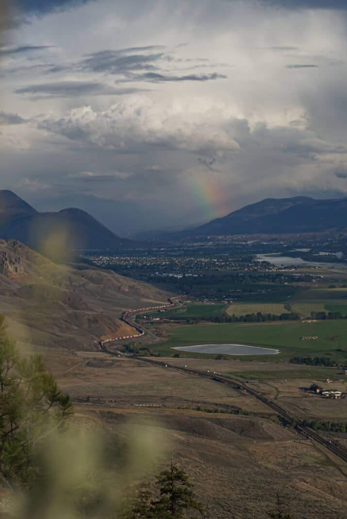 Vancouver to Kamloops road trip. Dry desert hills are seen to left with a train winding below in to Kamloops. A rainbow is also seen shining over Kamloops.