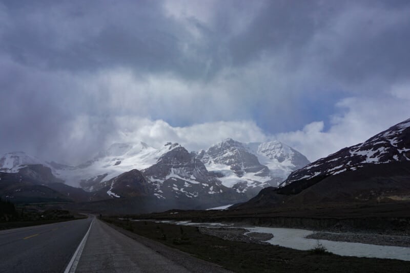 A view of the Icefields Parkway road running along the Athabasca River with looming dark snowy peaks in the background.